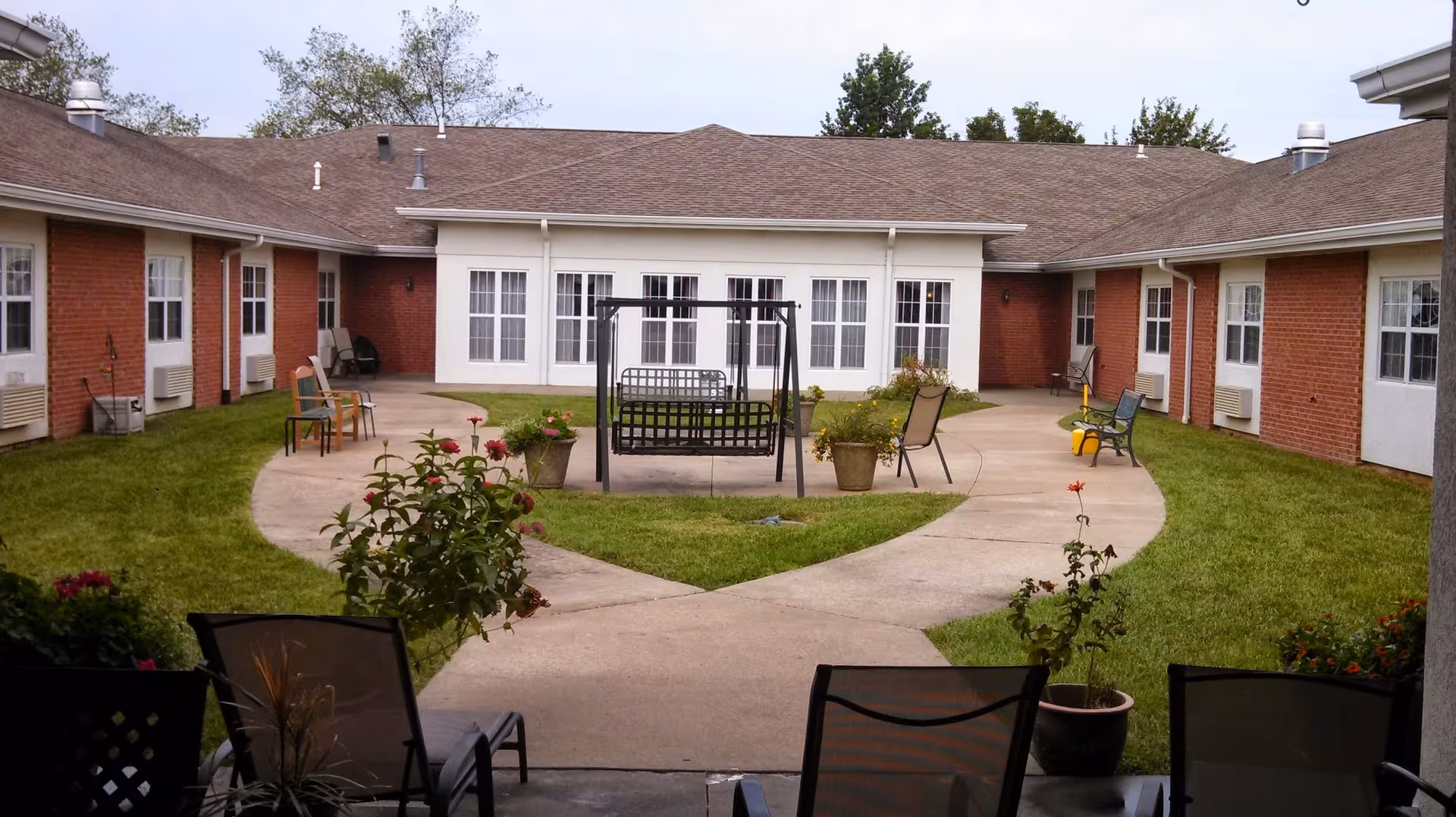 Outdoor courtyard area of a senior living facility with a concrete pathway forming a loop around a grassy area. There are several chairs and a black metal swing in the center of the courtyard. The building surrounding the courtyard has red brick walls and white-framed windows and doors.
