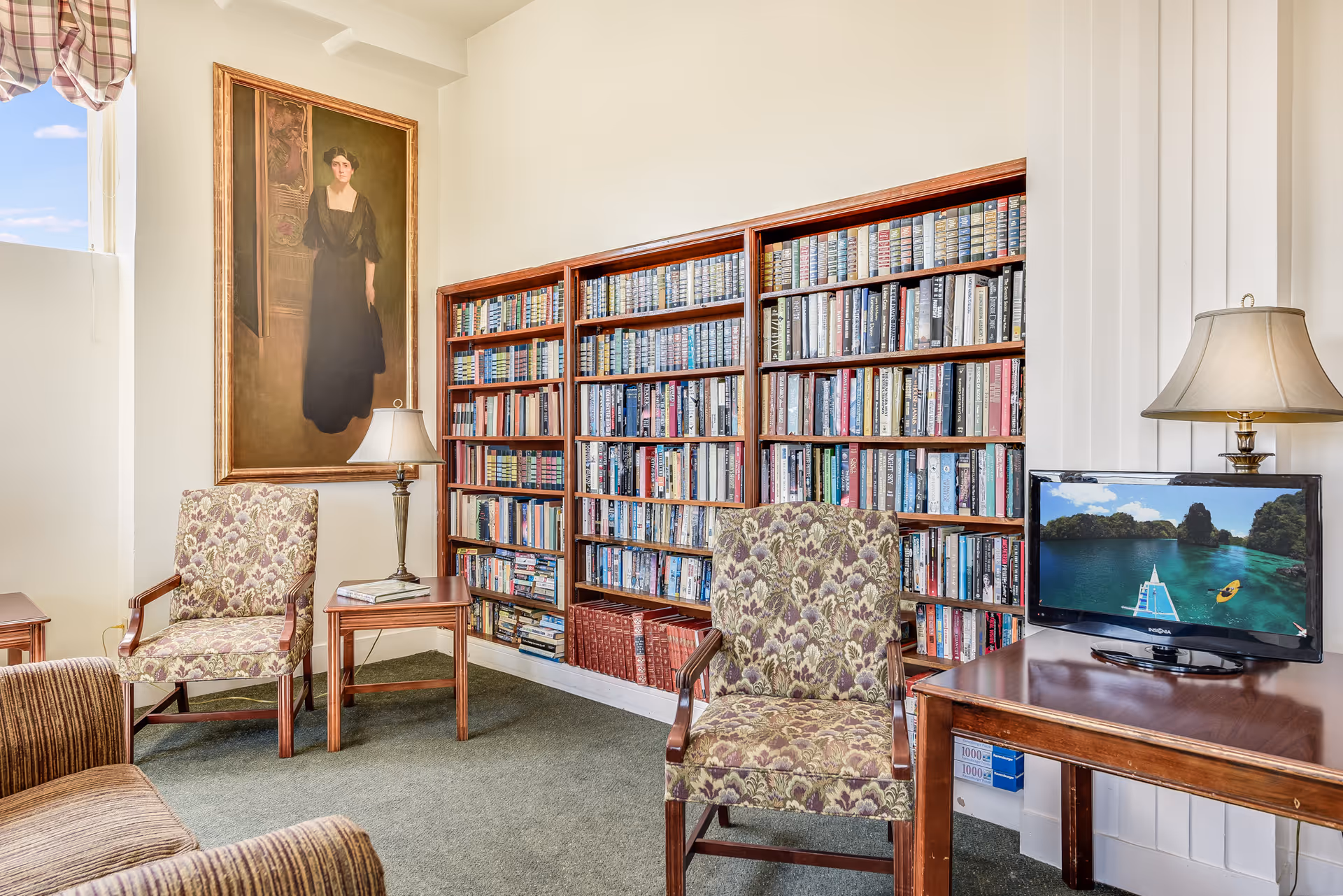 A cozy reading room with two floral upholstered armchairs, a wooden side table with a lamp and book, a large bookshelf filled with books, a small TV on a wooden table displaying a scenic water and island view, and a large portrait painting of a woman in a black dress hanging on the wall.