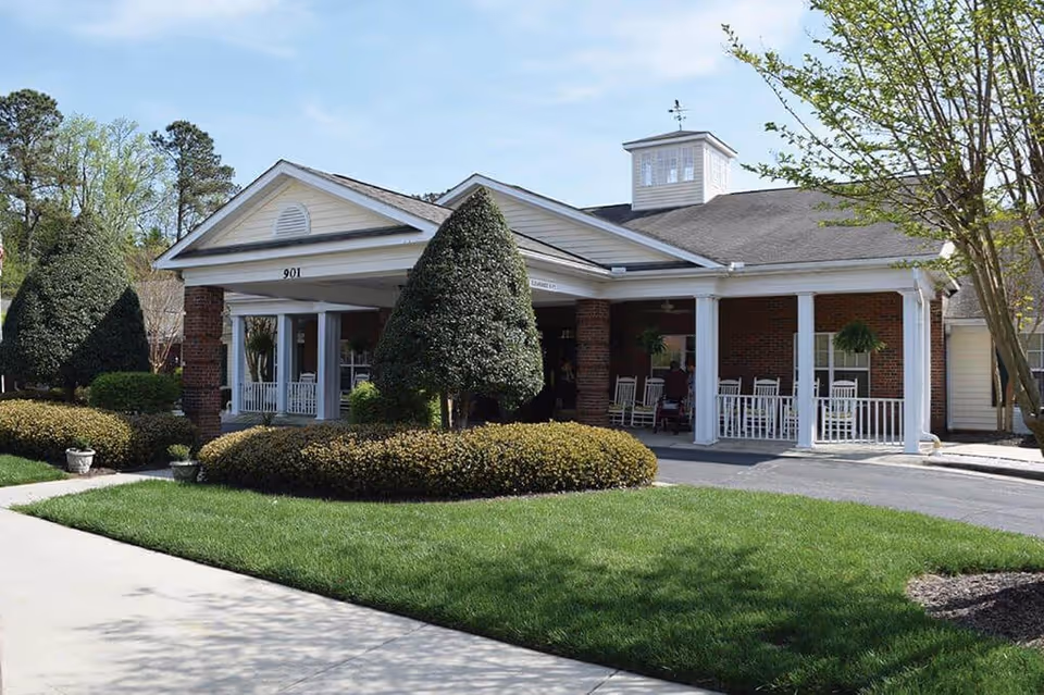 Exterior view of a single-story brick building with white columns and a covered porch area featuring rocking chairs. The building is surrounded by neatly trimmed bushes, shaped trees, and a well-maintained lawn under a clear sky.