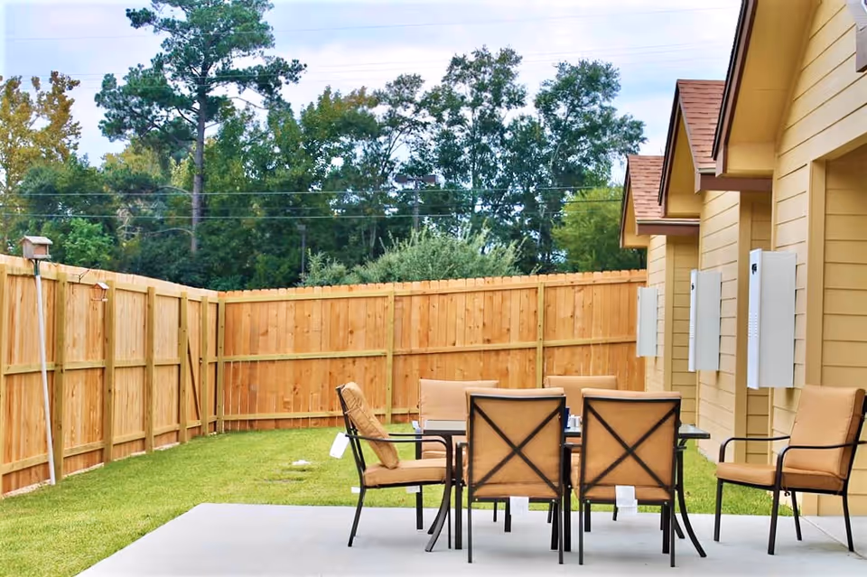 Outdoor patio area with a glass-top table and six cushioned chairs on a concrete slab, surrounded by a wooden privacy fence and adjacent to a yellow building with multiple doors and utility boxes. Trees and greenery are visible beyond the fence.