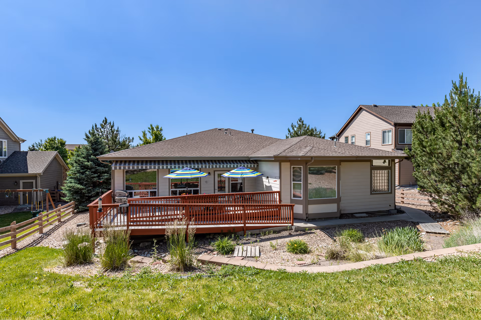 View of the back exterior of a single-story residential building with a wooden deck featuring two striped umbrellas. The yard has green grass, shrubs, and a wooden fence, with neighboring houses visible under a clear blue sky.