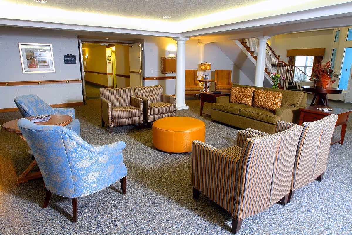 A spacious senior living facility lounge area with various seating options including two blue patterned armchairs, four striped armchairs, a green sofa with patterned cushions, and an orange round ottoman in the center. The room features carpeted flooring, white columns, a staircase, side tables with lamps, and floral arrangements near the entrance.
