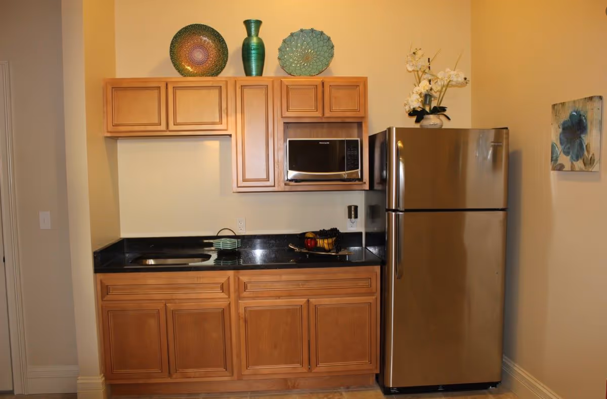 Small kitchenette with wooden cabinets, a black granite countertop, stainless steel refrigerator and microwave, and decorative plates and vase above the cabinets.