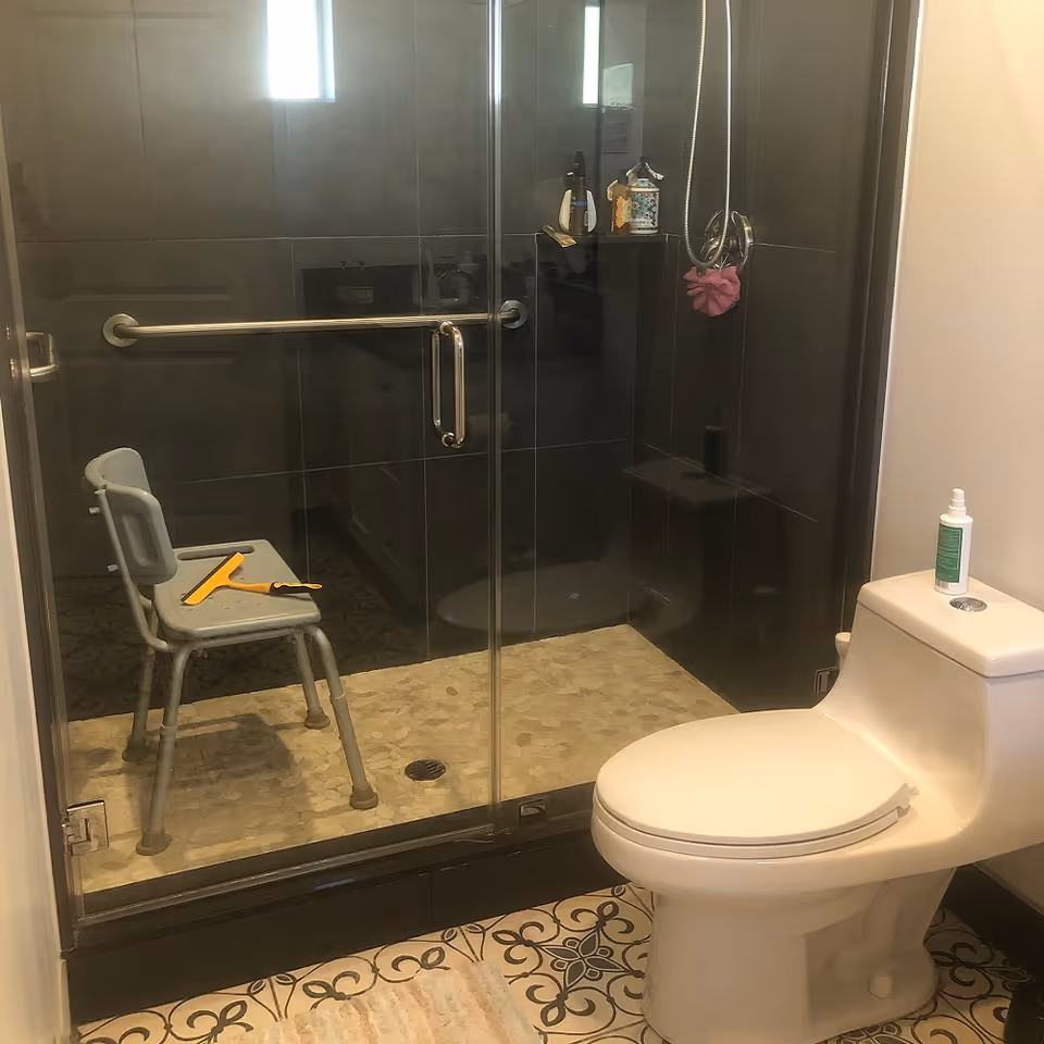 Bathroom with a glass-enclosed shower featuring a metal grab bar and a gray shower chair inside. A yellow squeegee is placed on the chair. The floor has patterned tiles, and there is a white toilet with a bottle of spray cleaner on top. A small window is visible above the shower area.