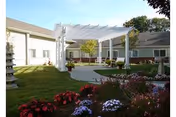 Outdoor courtyard with a white pergola, seating area, lawns and flower beds in front of a single-story building.