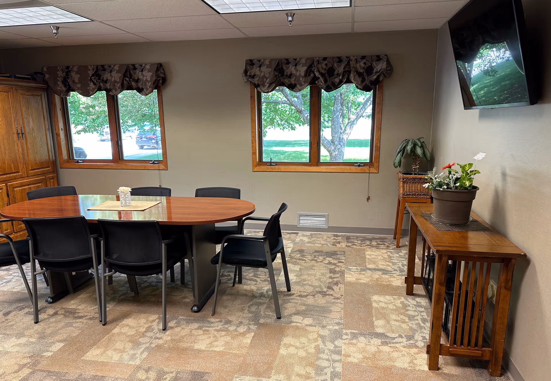 A meeting or dining room with a large oval wooden table surrounded by black chairs. There are two windows with patterned valances showing green trees outside. A wooden cabinet is on the left wall, and a wooden console table with potted plants is on the right wall beneath a mounted flat-screen TV. The floor is carpeted with a patterned design.