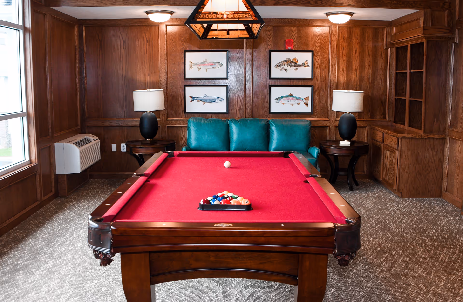 A cozy game room with a red felt pool table in the center, a teal leather couch against a wood-paneled wall, two side tables each with a black lamp, and four framed fish illustrations hanging on the wall above the couch.