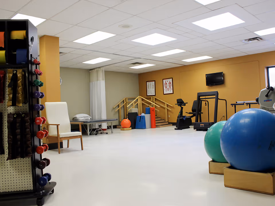 A spacious physical therapy or rehabilitation room with exercise equipment including stationary bikes, a treadmill, large exercise balls, dumbbells, resistance bands, and a therapy table. The room has a white floor, beige and gray walls, ceiling lights, and anatomical charts on the wall.