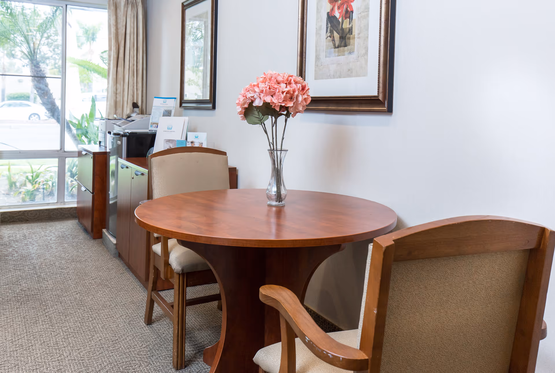 A small round wooden table with a glass vase holding pink flowers sits between two upholstered chairs in a room with a large window. There is a framed floral artwork on the white wall above the table, and a cabinet with brochures and a printer is visible near the window.