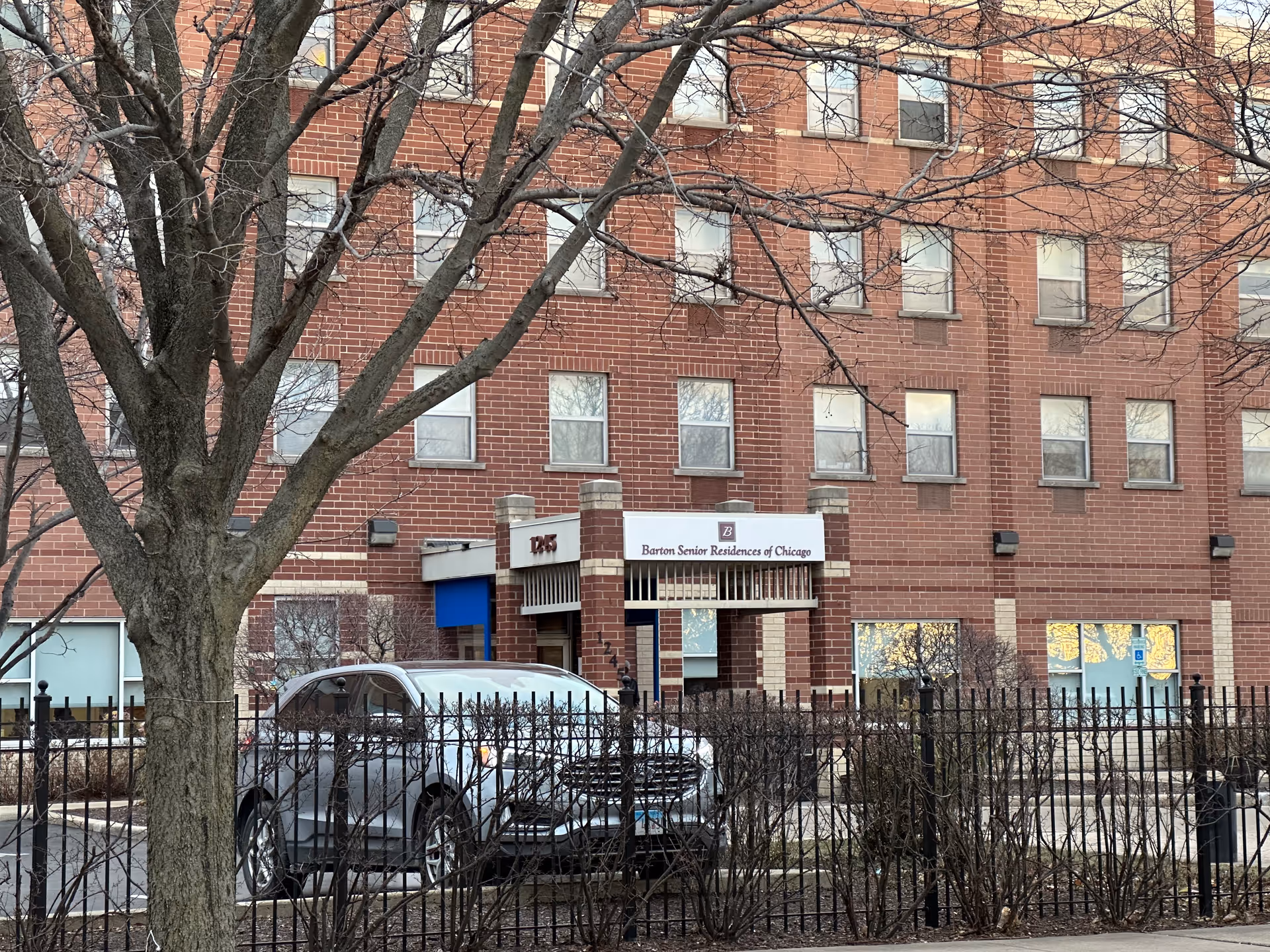Exterior view of Barton Senior Residences of Chicago, a multi-story brick building with several windows. A silver SUV is parked in front behind a black metal fence, and a large tree with bare branches is in the foreground.