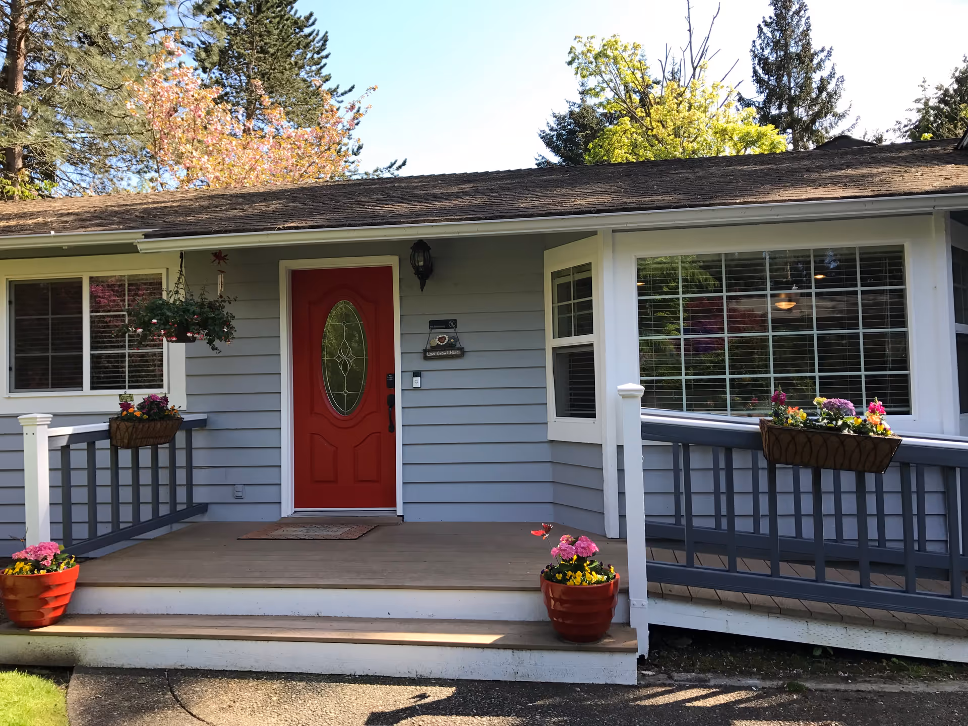 Front porch of a single-story house with a red front door, gray siding, and white trim. The porch has a wooden floor with steps leading up to it, decorated with flower pots containing colorful flowers. There is a ramp with railings on the right side of the porch, also adorned with flower boxes. Trees with green and pink foliage are visible in the background.