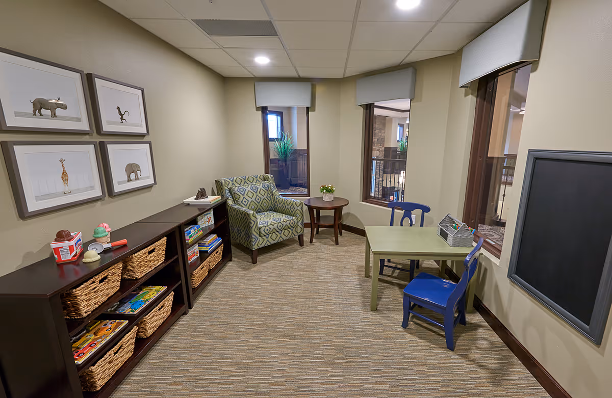 A cozy interior room with beige walls and carpeted floor featuring a small green table with two blue chairs, a patterned armchair next to a round wooden side table with a small plant, and two dark wooden bookshelves filled with baskets and children's books and toys. Four framed animal pictures hang on the wall, and there is a large blackboard on the opposite wall. Two windows with valances look out into another indoor area.