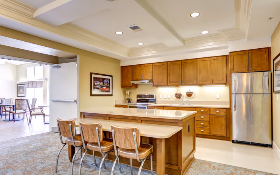 A bright kitchen area with wooden cabinets, a stainless steel refrigerator, stove, and a kitchen island with three wooden chairs. The kitchen opens to a dining area with tables and chairs visible through an open doorway.