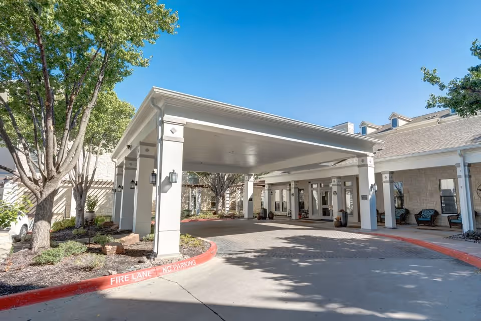Entrance area of Ridgmar Place senior living facility showing a covered drop-off zone with white pillars, outdoor seating, trees, and clear blue sky.