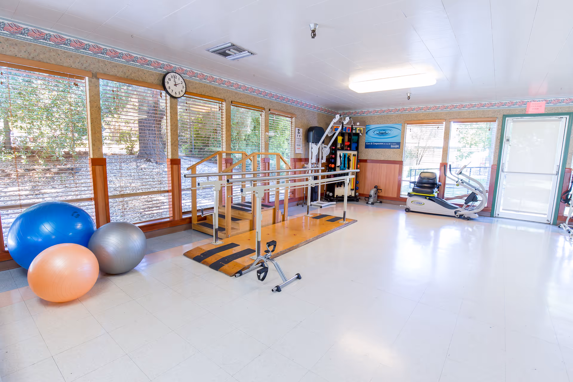 A bright rehabilitation or exercise room with large windows showing outdoor trees. The room contains exercise balls in blue, silver, and peach colors, a set of parallel bars with a small ramp and steps for physical therapy, a stationary recumbent exercise bike, and various exercise equipment on shelves. The walls have a decorative border near the ceiling, and a clock is mounted above the windows.