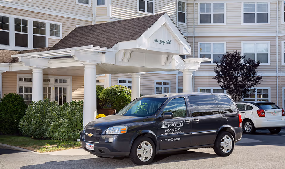 Exterior view of Benchmark Senior Living at Forge Hill showing the entrance with white pillars and a covered driveway. A black van with the facility's name, phone number, and website is parked in front. The building has beige siding and multiple windows, with some greenery and bushes around the entrance.