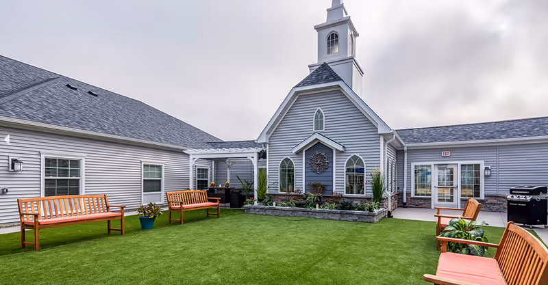A well-maintained courtyard with artificial turf, wooden benches, planters and a grey building with a small steeple.
