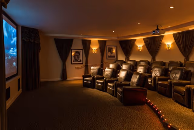 A cozy home theater room with two rows of black leather recliner chairs facing a large screen displaying a movie scene. The room is dimly lit with wall sconces and has dark curtains covering the windows. Framed pictures hang on the walls behind the chairs.