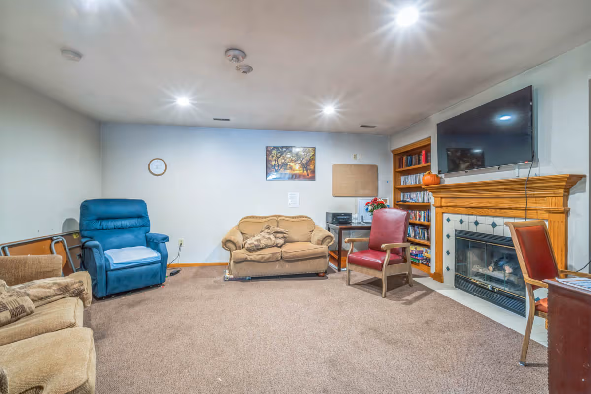 A cozy living room with beige carpet and neutral walls. The room features a blue recliner chair, a beige loveseat with a blanket, two red armchairs, a wooden bookshelf filled with books, and a wooden fireplace mantel with a flat-screen TV mounted above it. There is a framed picture on the wall and a small table with a printer and a flower pot beside the loveseat.
