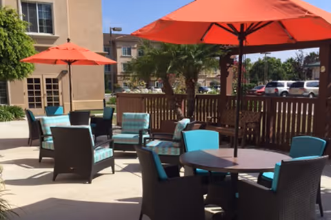 Outdoor patio area with several round tables and chairs featuring turquoise cushions. Large orange umbrellas provide shade over the seating. A wooden fence encloses the patio, with a building and parked cars visible in the background under a clear blue sky.