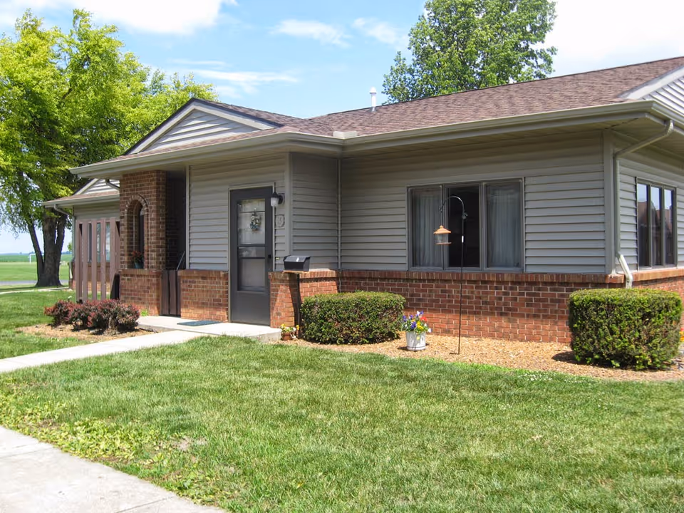 Exterior view of a single-story residential building with gray siding and red brick accents. The building has a brown shingled roof, a front door with a small porch, and several windows. There are neatly trimmed bushes and a green lawn in front, with a tree on the left side and a clear blue sky above.