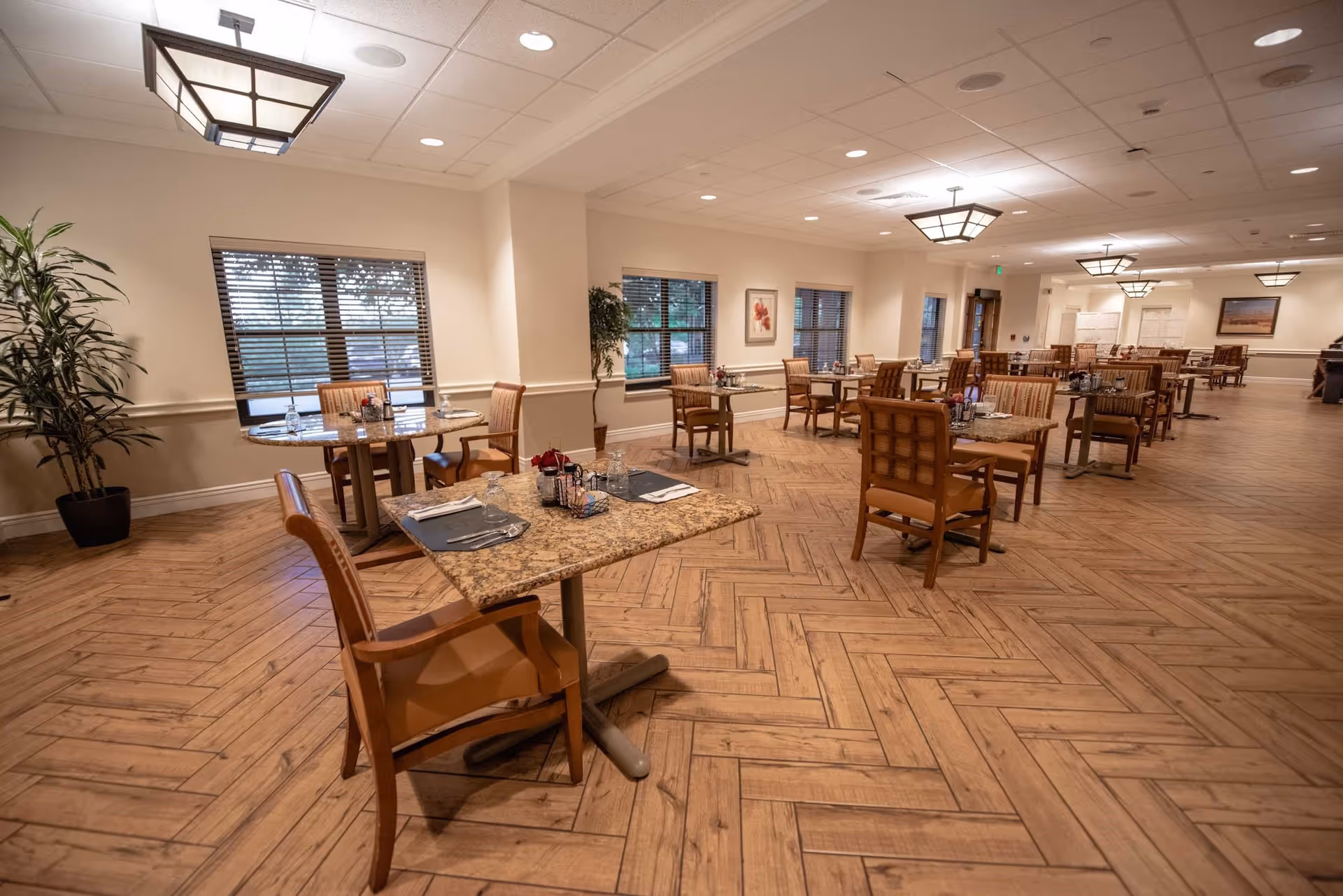 A spacious dining room with multiple tables and chairs arranged neatly. The room has wooden flooring with a herringbone pattern, large windows with blinds, potted plants, framed artwork on the walls, and ceiling lights providing warm illumination.
