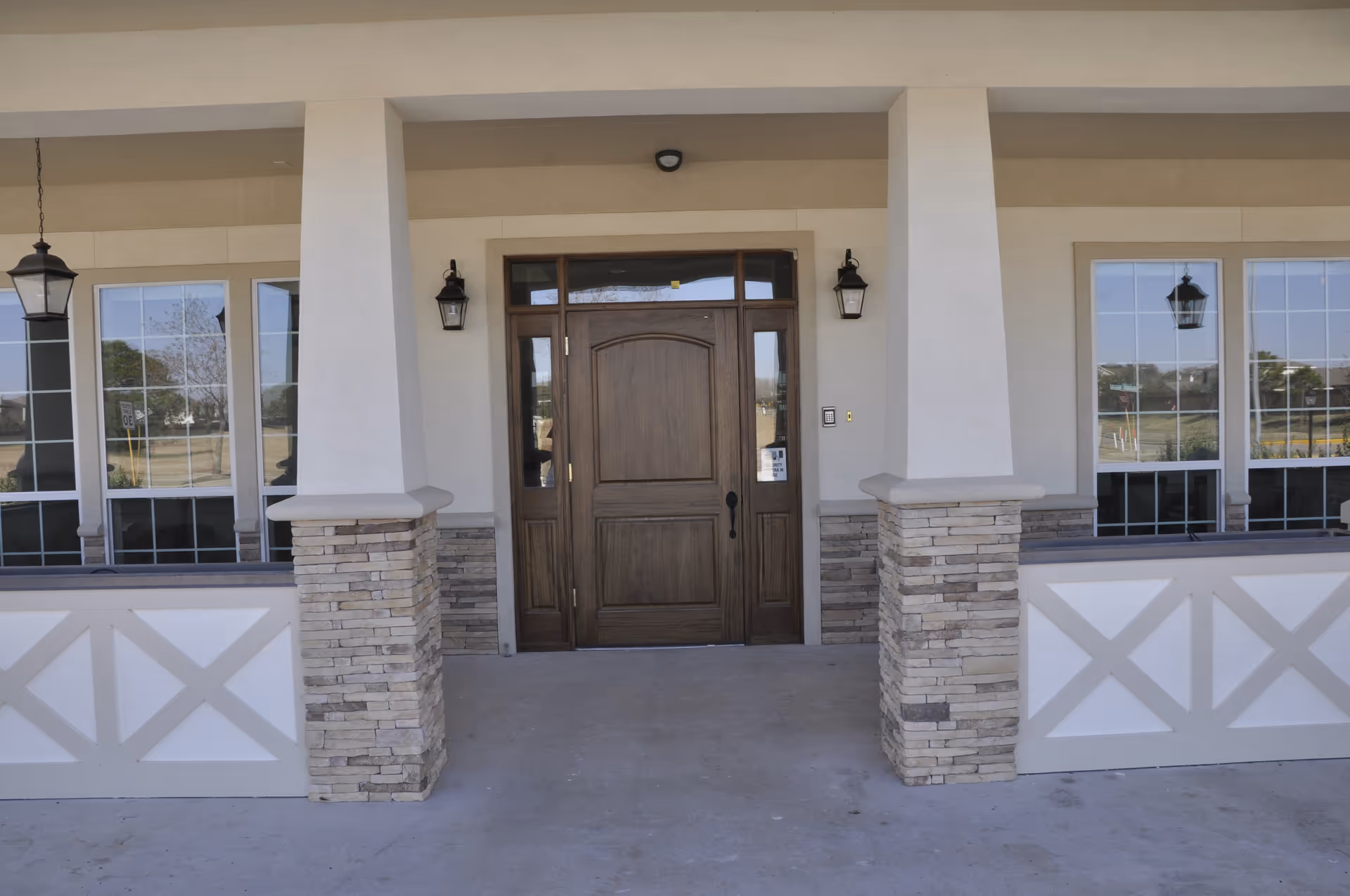 Front entrance of a building with a wooden door flanked by stone-clad columns, windows, and porch lanterns.