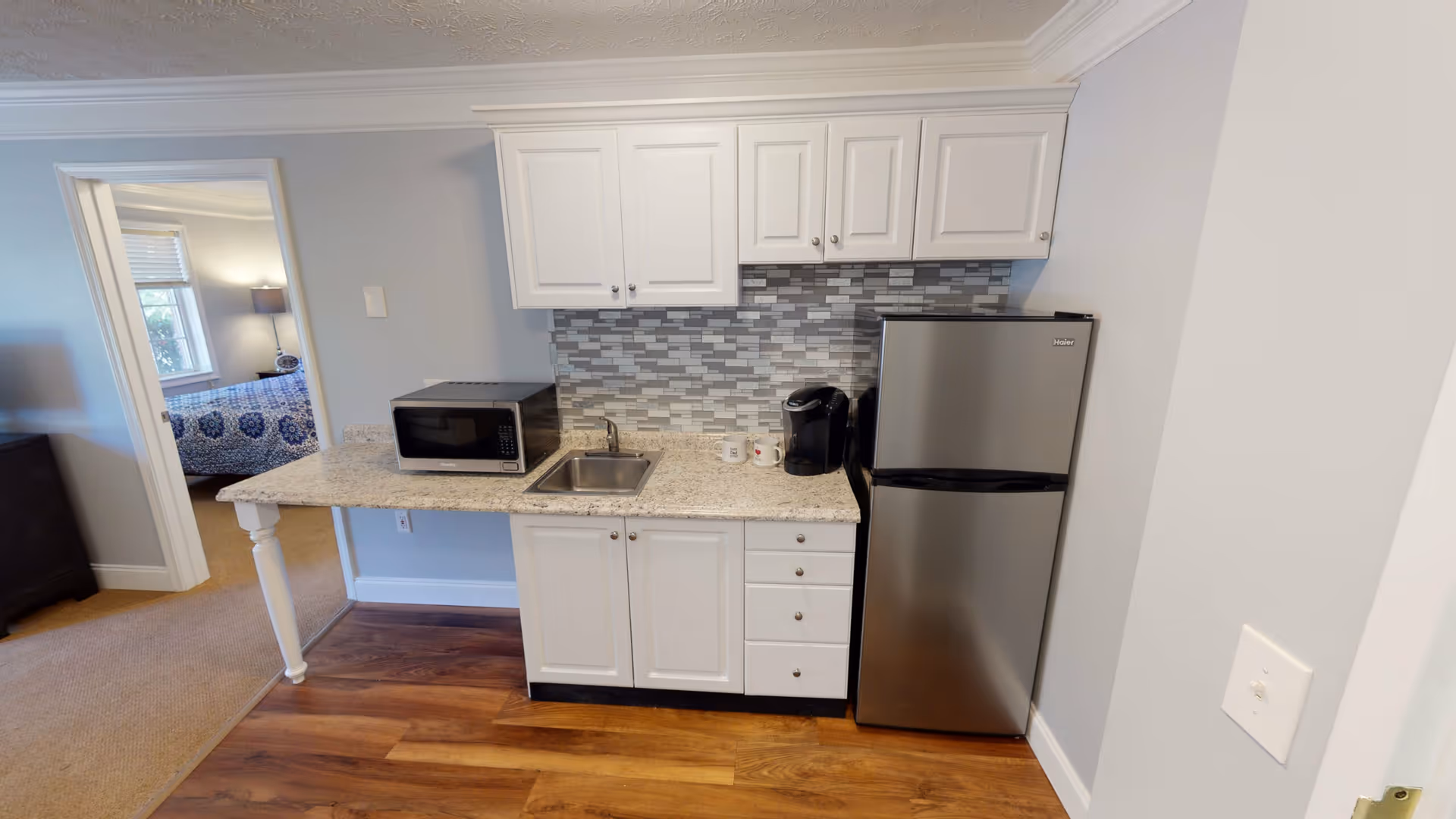 Compact kitchenette with white cabinets, granite countertop, sink, microwave, coffee maker and stainless steel refrigerator, with a bedroom visible through a doorway.