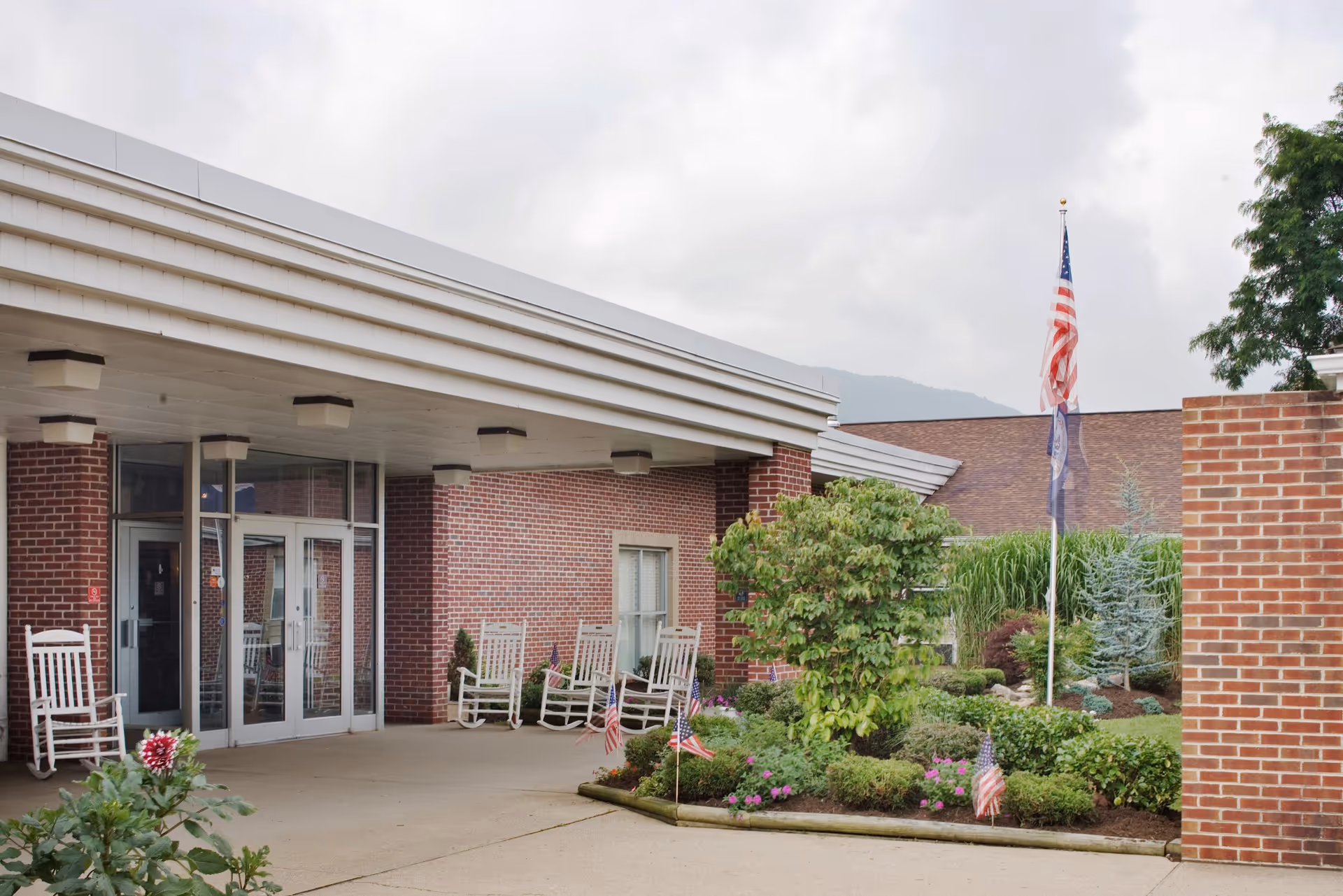 Entrance of a brick building with a covered walkway, several white rocking chairs, and a landscaped garden area with small American flags and a flagpole displaying the American flag and another flag. The sky is cloudy and there are mountains visible in the background.