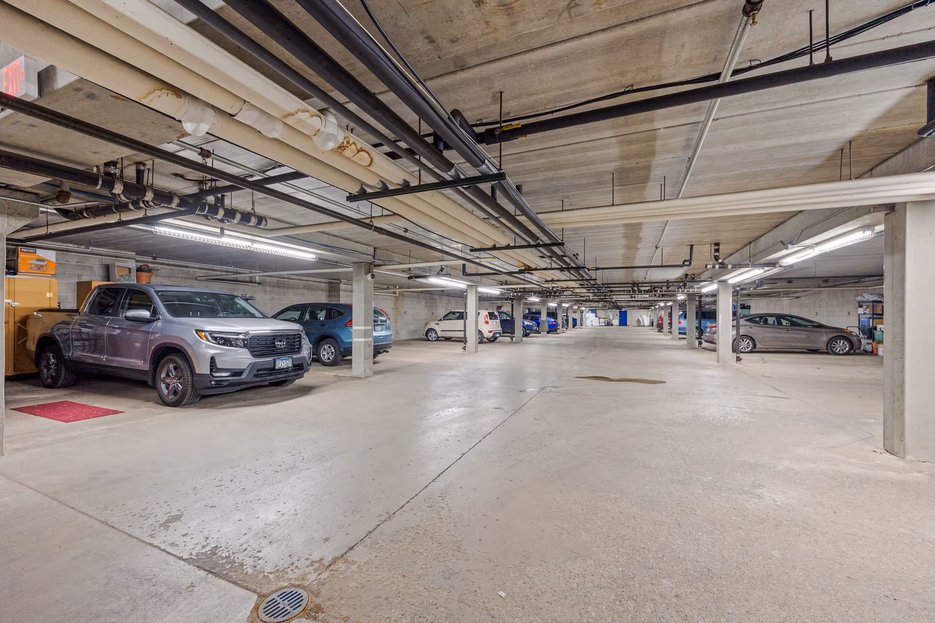 Underground concrete parking garage with parked cars, exposed ceiling pipes, and support columns.