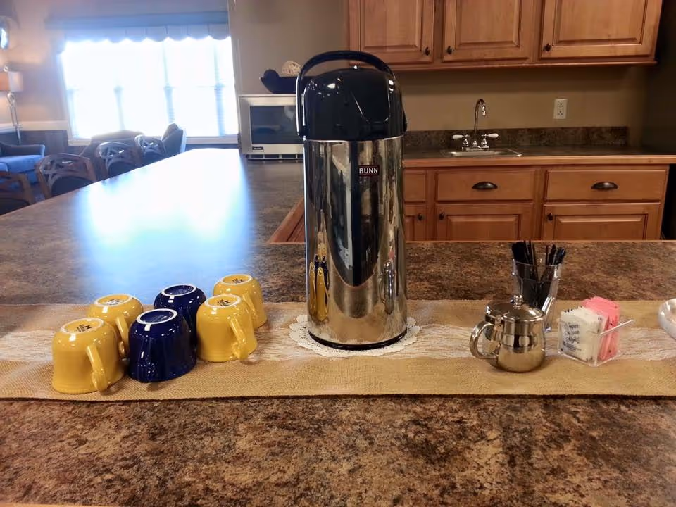 A kitchen counter with a silver Bunn coffee dispenser in the center, surrounded by six upside-down coffee mugs in yellow and blue. To the right of the dispenser are a small silver creamer pitcher, a container with sugar packets, and a cup holding black stir sticks. In the background, there are wooden cabinets, a sink, a microwave, and a seating area with chairs near a window.