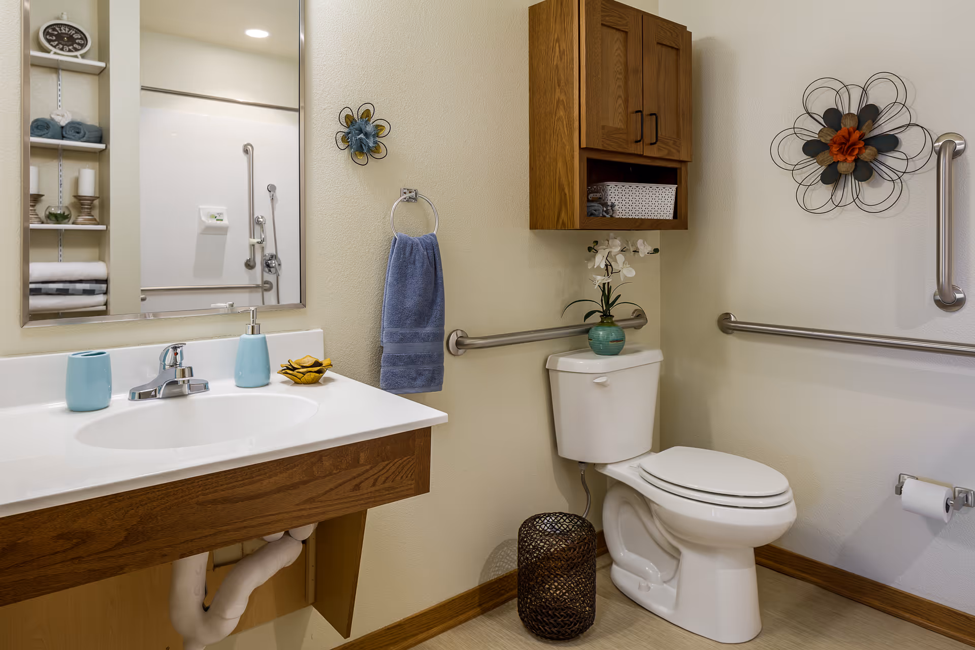 A clean and accessible bathroom featuring a white toilet with a wooden cabinet above it, a white sink with a wooden vanity, a mirror, and a blue hand towel hanging on a ring. The bathroom has safety grab bars on the walls and decorative wall art, with shelves holding towels and candles visible in the mirror reflection.