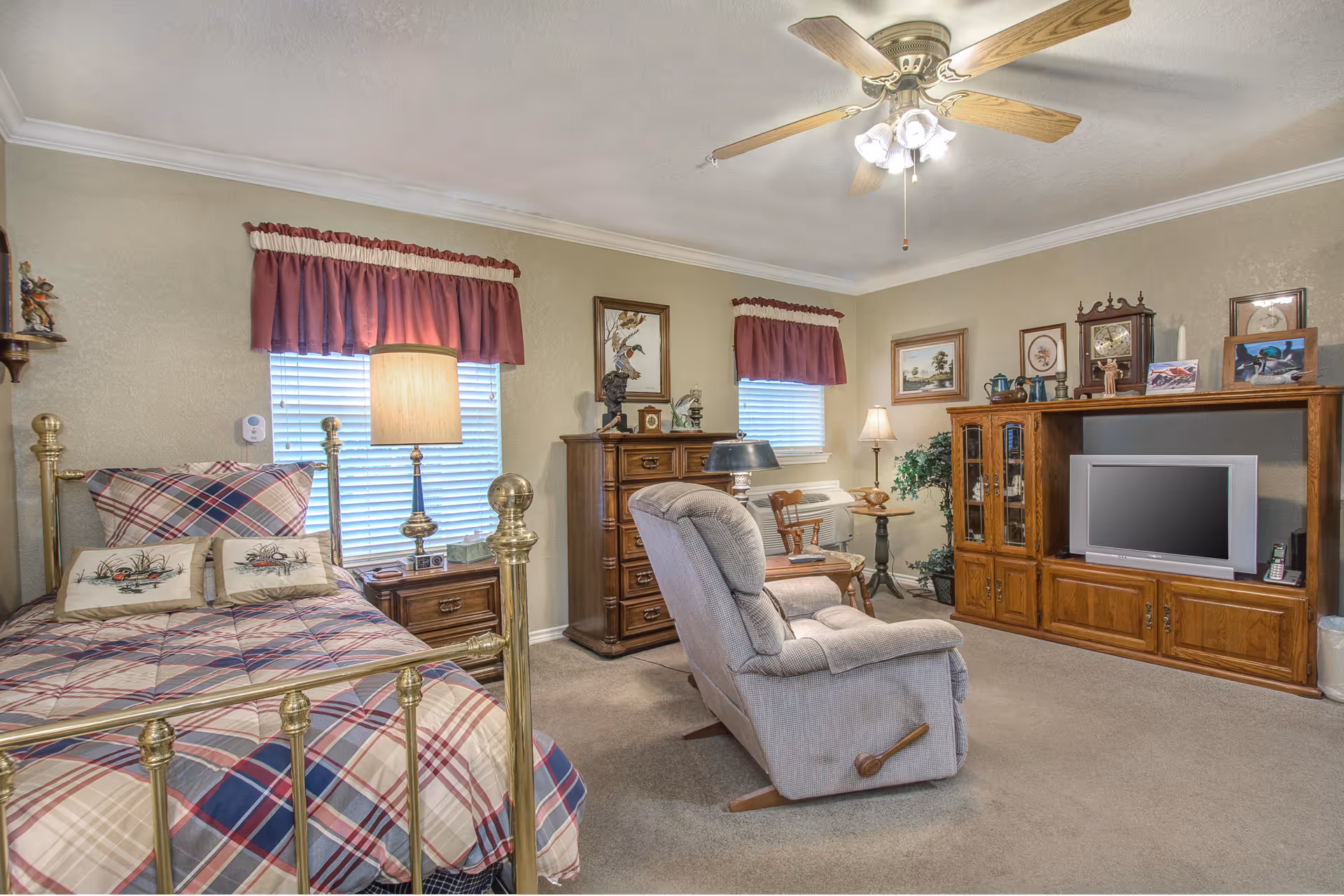 A cozy bedroom with a brass bed frame dressed in plaid bedding and decorative pillows. The room features two windows with red valances, a wooden dresser, a reclining armchair, a wooden entertainment center with a TV, and various framed pictures and decorative items. The ceiling has a fan with lights, and the walls are painted beige.
