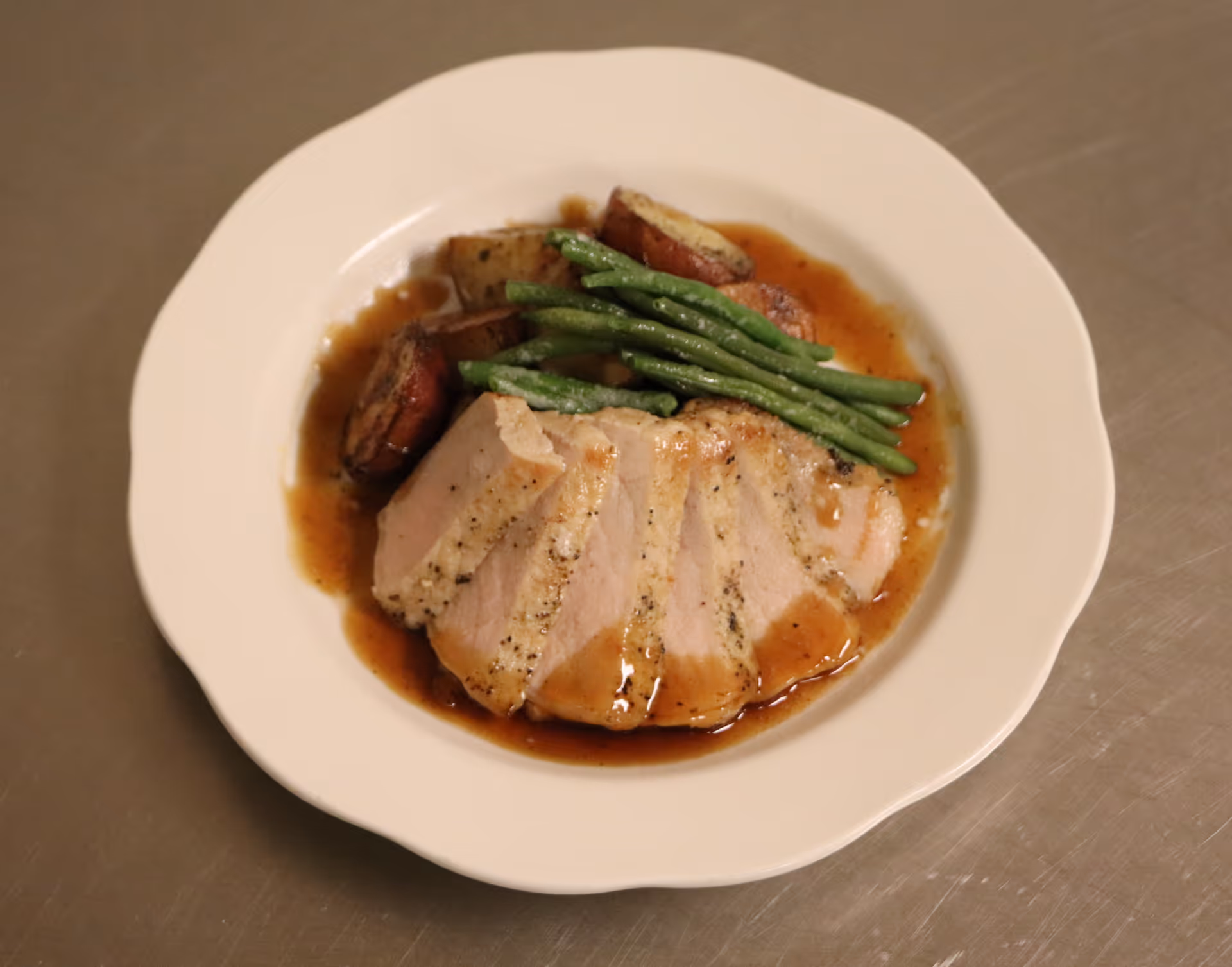 A white plate with sliced roasted pork, green beans, and roasted potatoes, all served with brown gravy on a stainless steel surface.