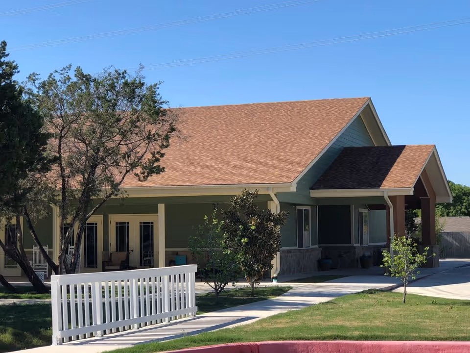 Single-story green building with a gabled brown roof, front porch, white railing, and surrounding lawn and trees.