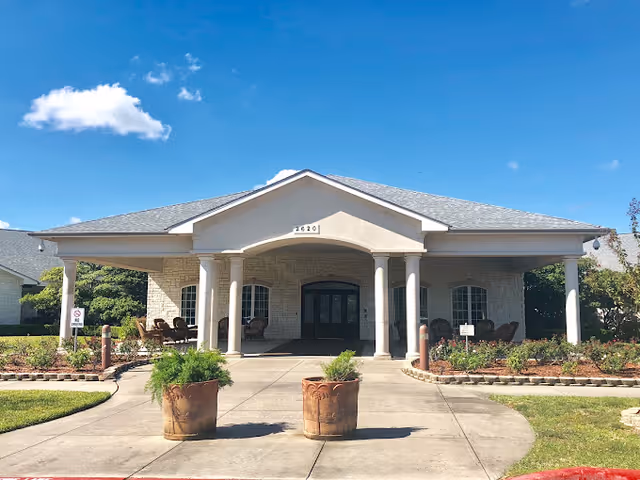 Front exterior view of a single-story building with a covered entrance supported by four columns. The building has light-colored brick walls, a gray shingled roof, and the number 2920 displayed above the entrance. There are two large planters with greenery on the walkway leading to the entrance, and outdoor seating is visible on the porch area. The sky is clear with a few clouds.