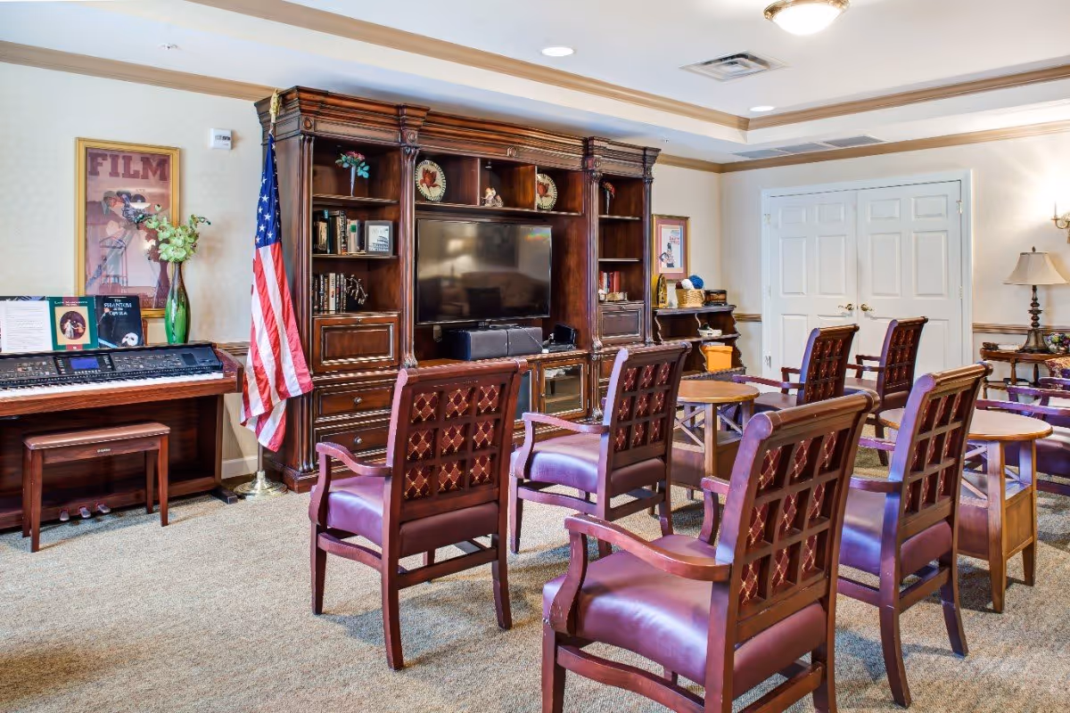 Well-lit common activity room with rows of wooden chairs facing a large entertainment center and TV, a piano, and an American flag.