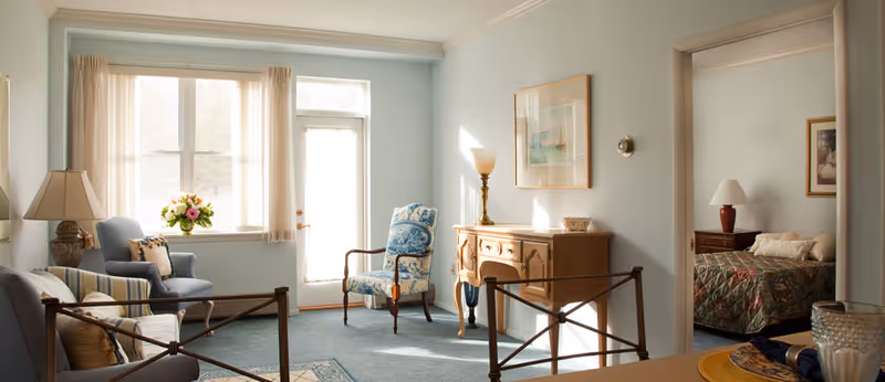 Sunlit living room with upholstered armchairs, a wooden console table, and a doorway opening into a bedroom.