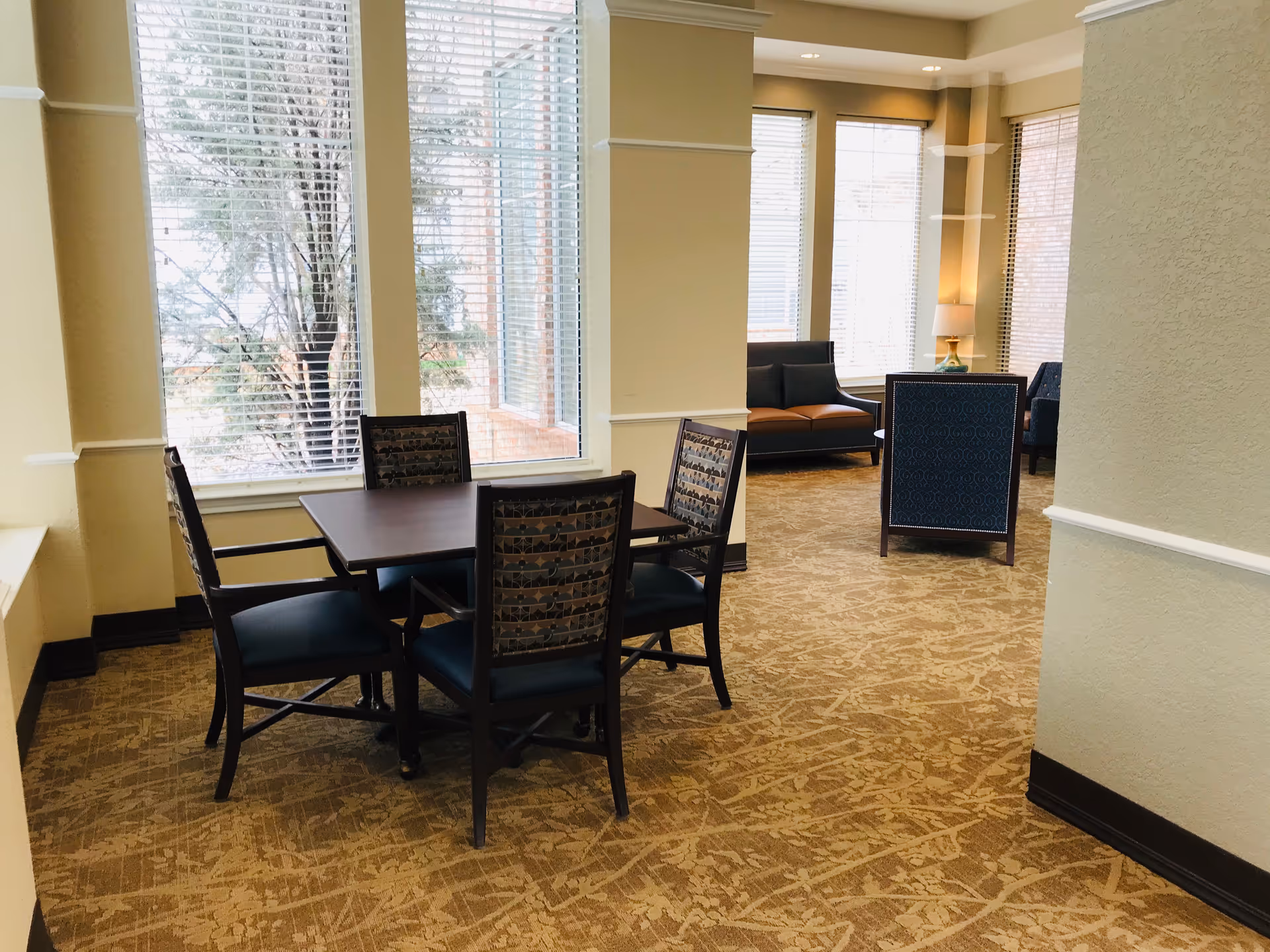 Sunlit common area with a square table and four chairs by large windows and lounge seating in the background.