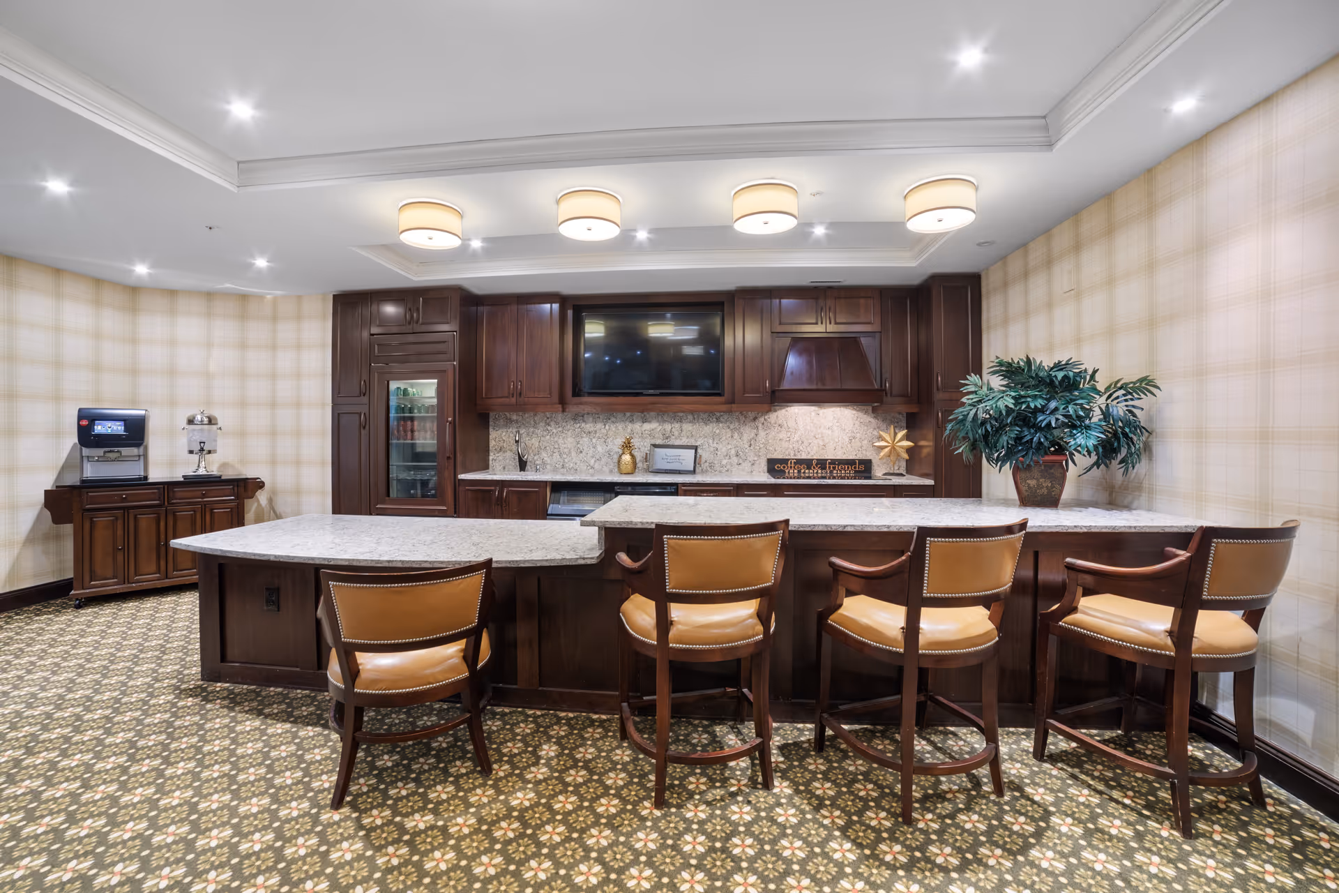A communal kitchen/bar area with a marble countertop, five leather bar stools, dark wood cabinets, and patterned carpet.