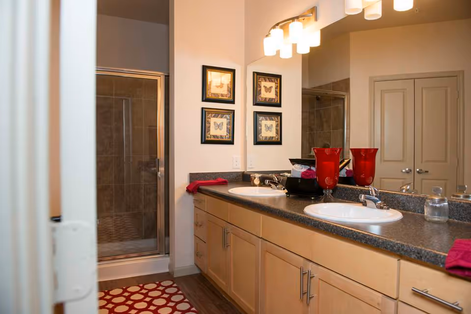 A modern bathroom with a double-sink vanity, large mirror, glass-enclosed shower, and red decorative accents.