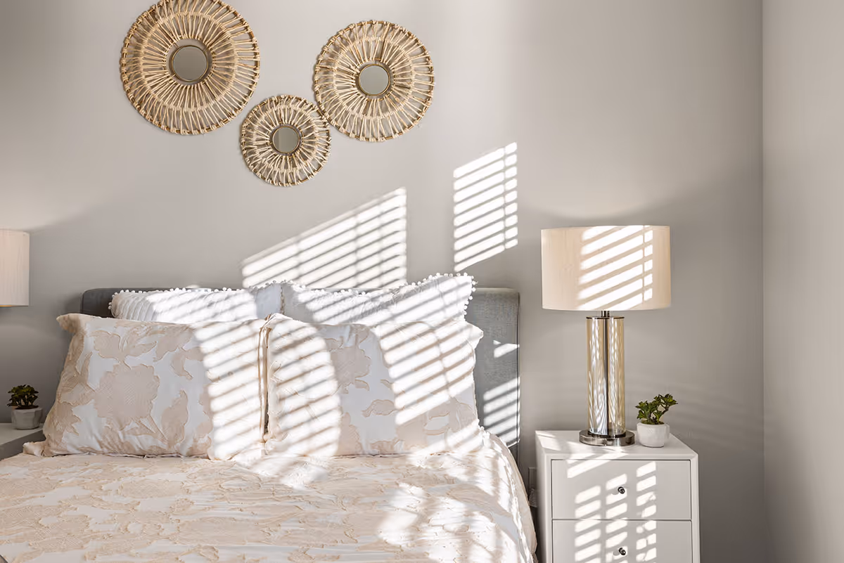 Sunlit bedroom featuring a bed with patterned pillows, three round woven mirrors above, and a nightstand with a lamp and small plant.