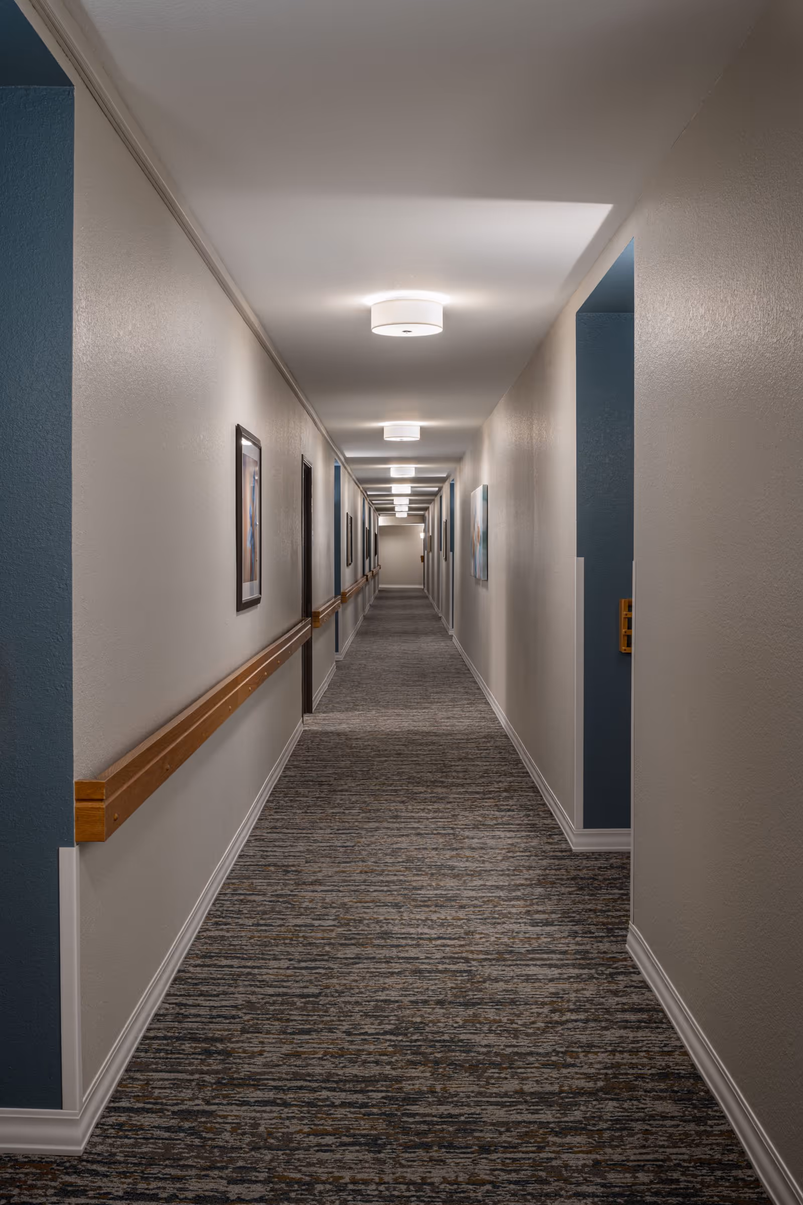 A long, well-lit hallway in a senior living facility with beige walls, carpeted floor, wooden handrails on the left side, and framed pictures hanging on the walls.