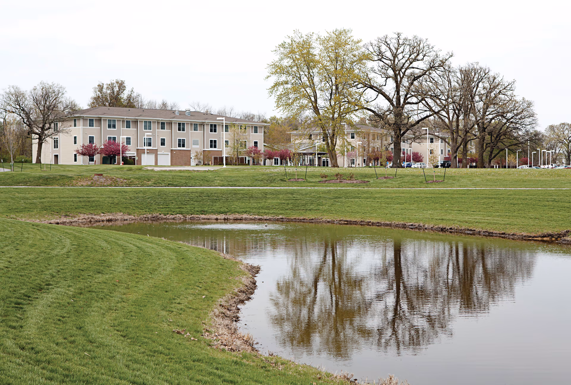 View of a senior living facility building named Timberland Village Senior Living with a large grassy area and a pond in the foreground. Trees with some autumn foliage surround the building under an overcast sky.