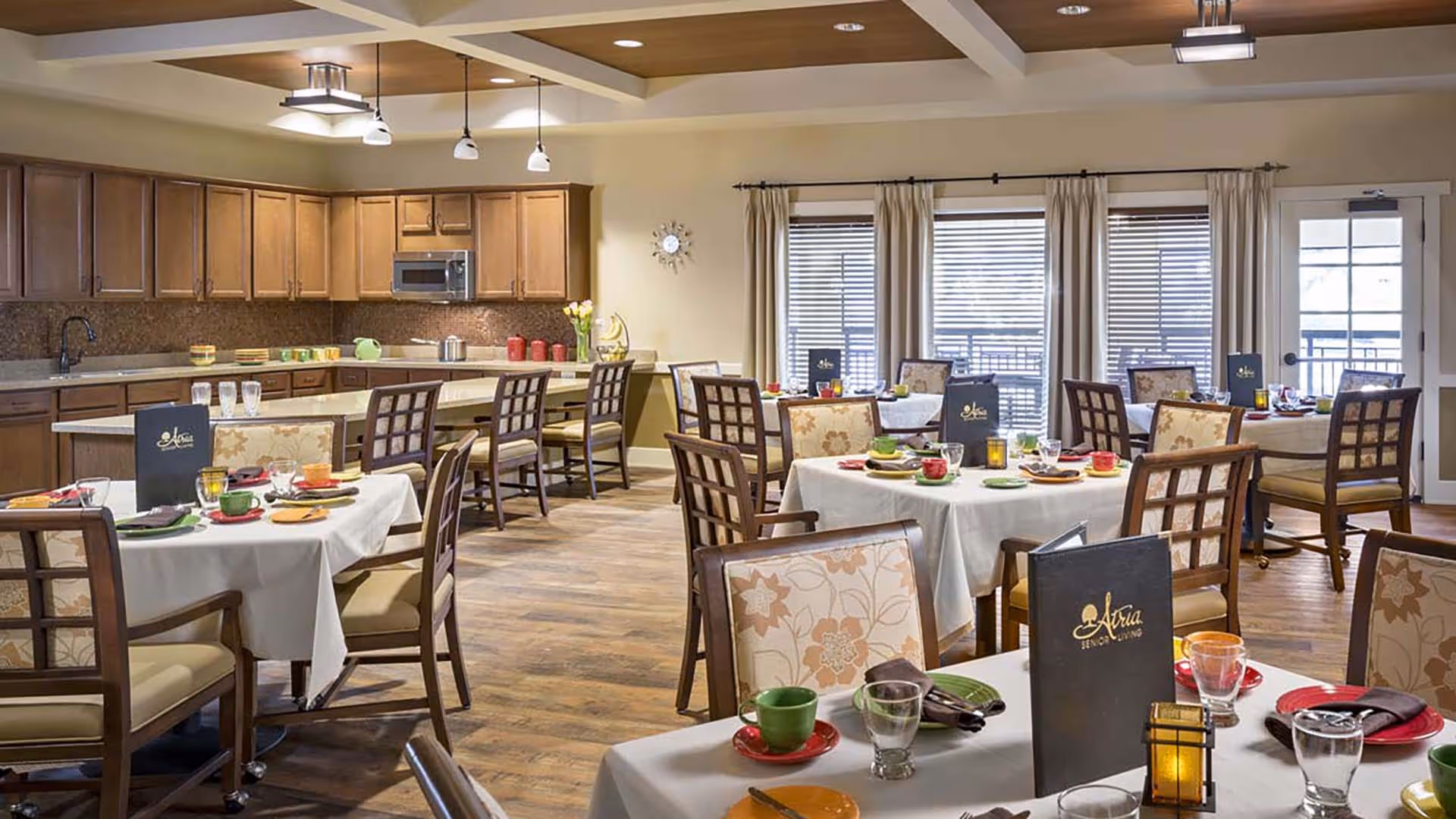A well-lit dining room in a senior living facility with several tables covered in white tablecloths, each set with colorful plates, cups, and glasses. The room features wooden chairs with floral upholstery, a kitchen area with wooden cabinets and a countertop with bar stools, and large windows with blinds and curtains letting in natural light.