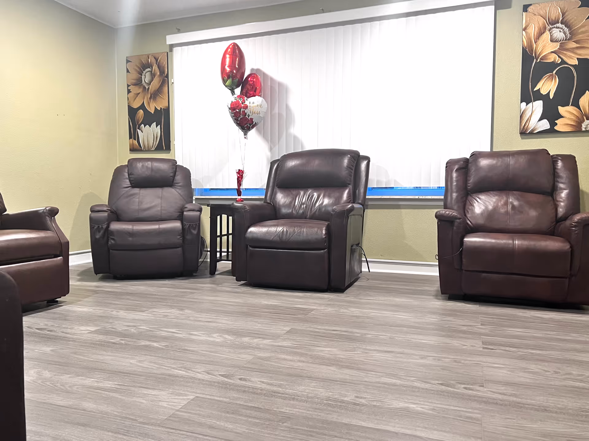 A room with four dark brown leather recliner chairs arranged around a small wooden side table. On the table, there are three balloons, two red and one white with red roses and the words 'I Love You'. The room has light green walls, two floral paintings on either side of a large window with white vertical blinds, and a light wood floor.