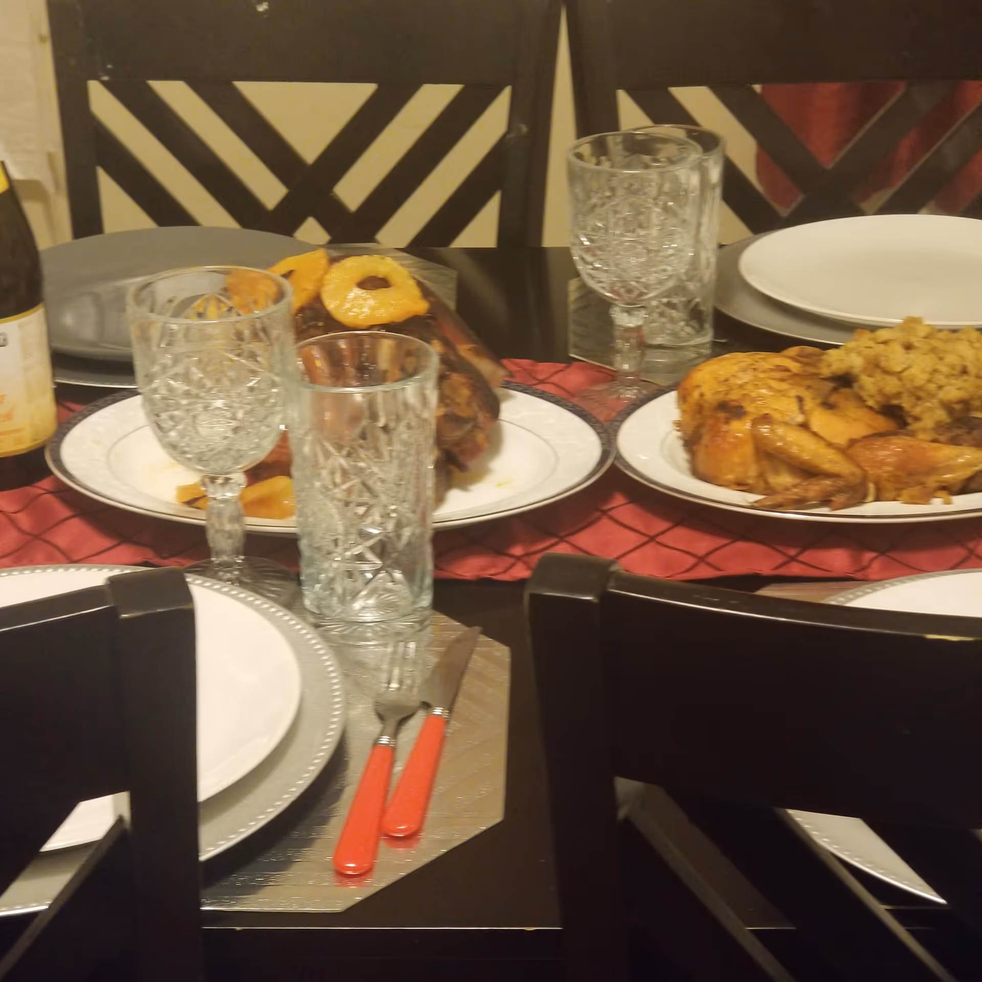 A dining table set for a meal with plates, crystal glasses, and silverware. The table has a red table runner and is adorned with cooked dishes including a roasted chicken with stuffing and a glazed ham garnished with pineapple rings. The chairs have a black and white geometric pattern on the backrests.