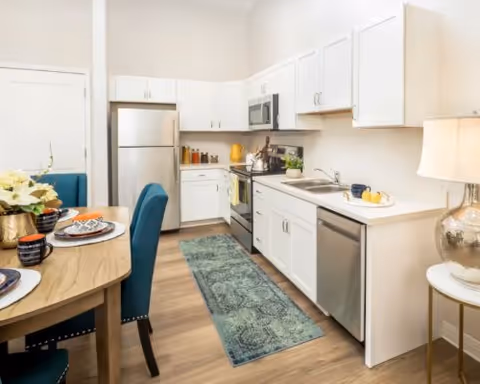 A modern kitchen and dining area featuring white cabinets, stainless steel appliances including a refrigerator, stove, microwave, and dishwasher. A wooden dining table with blue upholstered chairs is set with plates and mugs. A decorative rug runs along the floor in front of the kitchen counters. A lamp on a small round table adds warm lighting to the space.