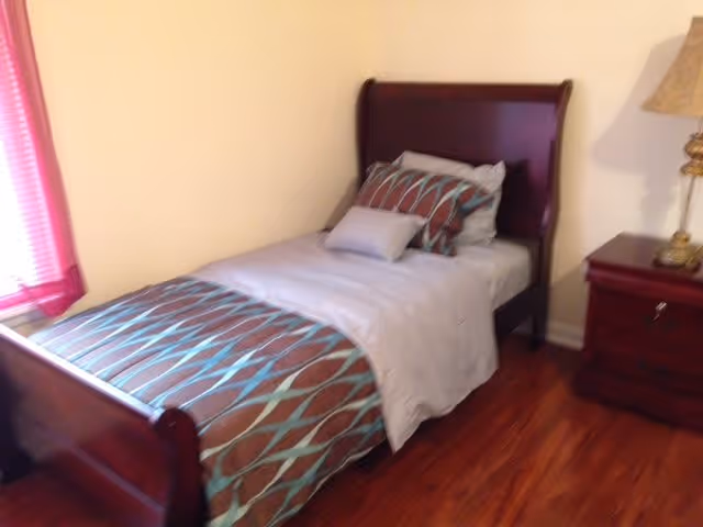 A small bedroom with a single wooden bed featuring a headboard and footboard. The bed is made with white sheets, a brown and blue patterned blanket, and matching pillows. Next to the bed is a wooden nightstand with a decorative lamp. The floor is wooden, and there is a window with pink blinds partially visible on the left side.