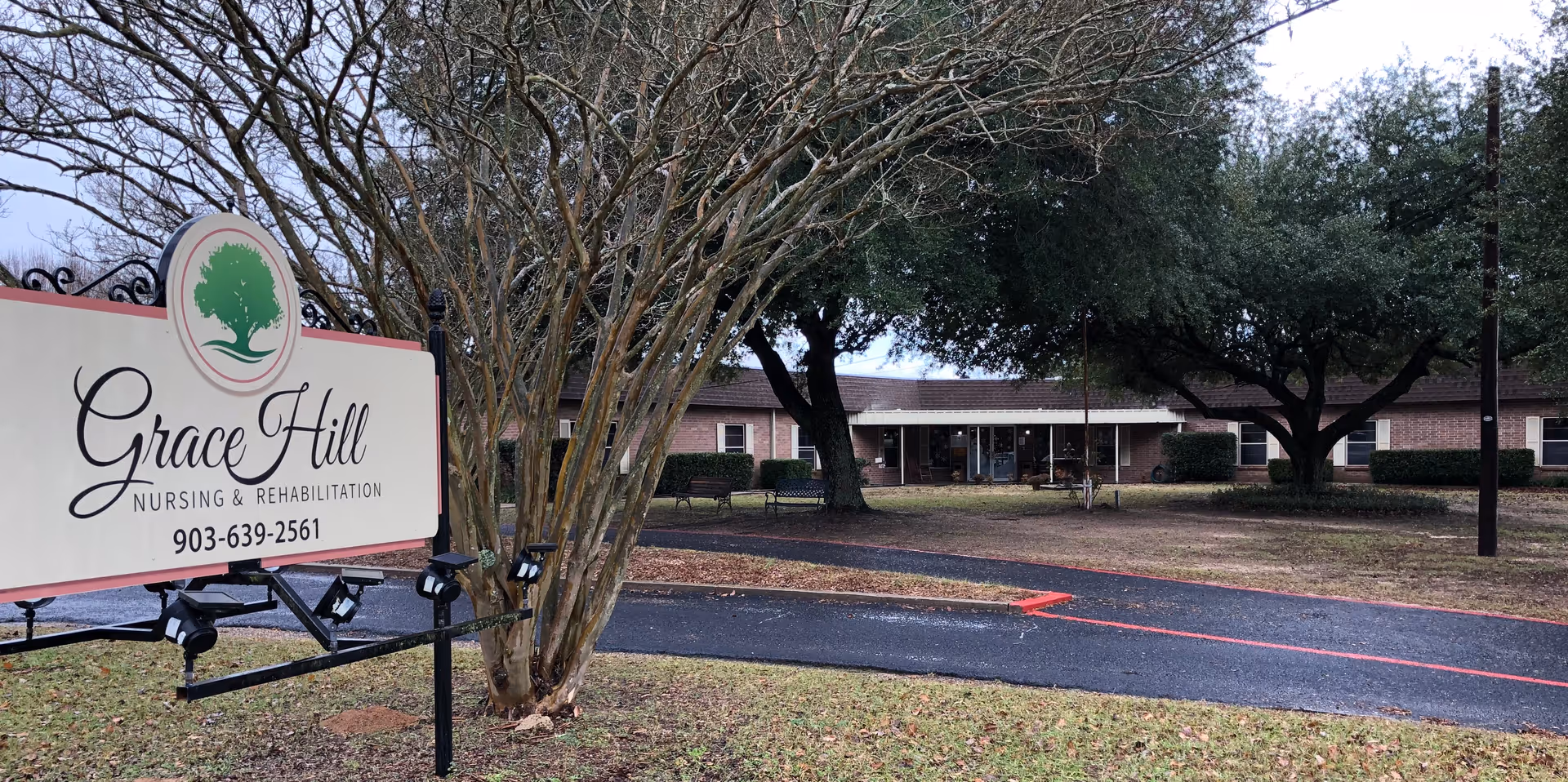 Outdoor view of a nursing and rehabilitation facility named Grace Hill, showing a sign with the facility's name and phone number, a paved driveway, trees without leaves, and a single-story brick building in the background.